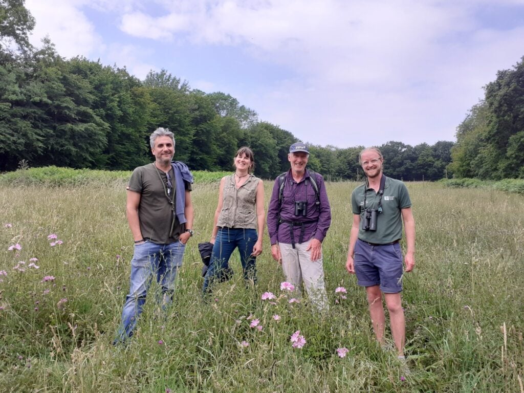 Four people smiling to camera standing in grassy field backed by woodland and bright but cloudy skies