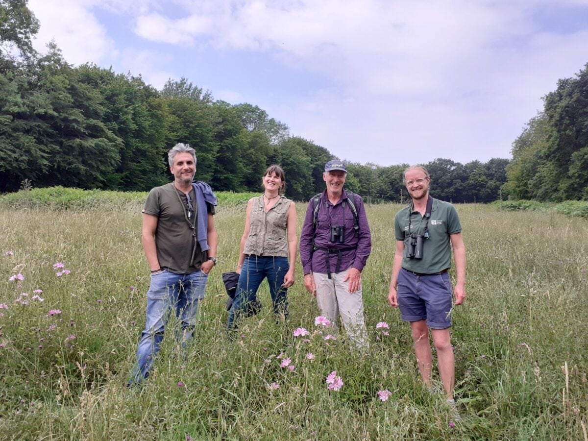 Four people smiling to camera standing in grassy field backed by woodland and bright but cloudy skies