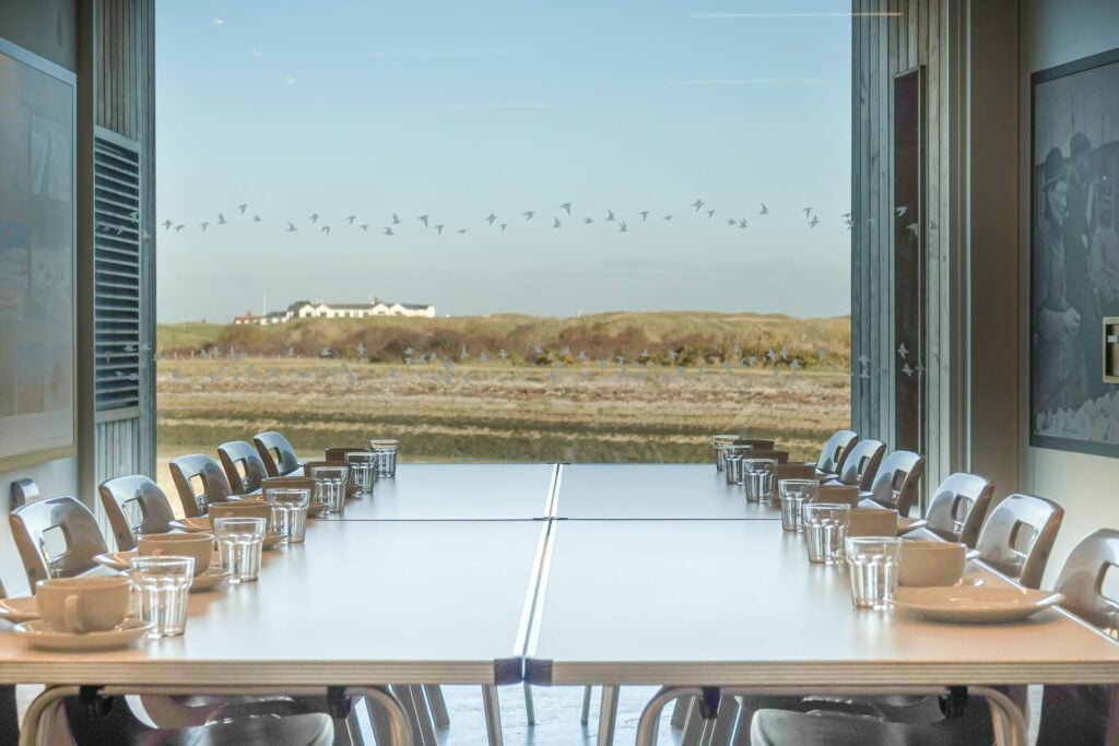 Table laid with glasses, cups and plates in front of big window showing greenery, white sided building in distance and low hills