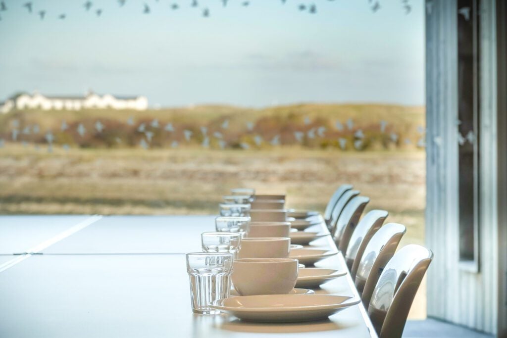 Chairs and table laid with cups, glasses and plates in front of large window with view over saltmarsh and sand dunes  
